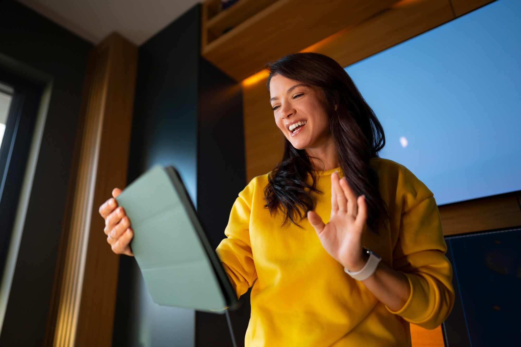 A woman in a yellow sweater laughing and waving during a video call on her tablet